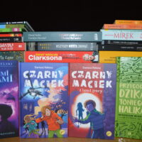 Na tle stosu książek ustawione książki dla dzieci i młodzieży. Widoczne tytuły: "Za niebieskimi drzwiami", "Czarny Maciek. Wenecki starodruk", "Czarny Maciek i tunel grozy", "Przygoda dzika Toniego Halika".