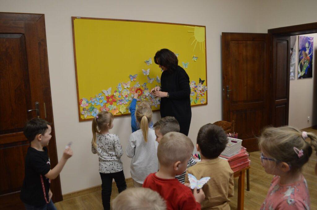 Zajęcia z przedszkolakami, grupa dzieci i osoba dorosła, w pomieszczeniu, przy kolorowej tablicy