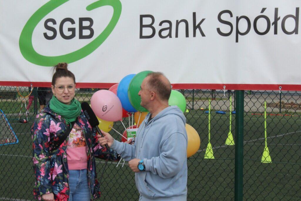 dwie osoby kobieta mężczyzna z mikrofonem przeprowadza wywiad na świeżym powietrzu, na boisku sportowym nad nimi napis SGB Bank spółdzielczy