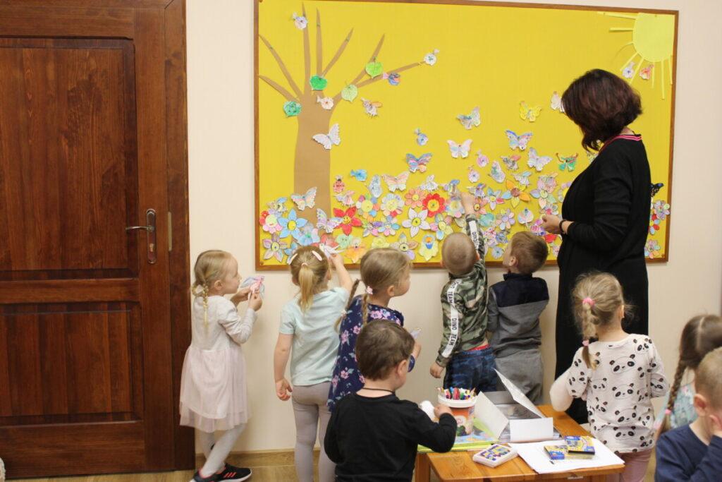 Zajęcia z przedszkolakami, grupa dzieci i osoba dorosła, w pomieszczeniu, przy kolorowej tablicy