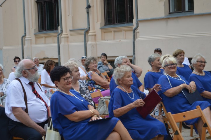 Na krzesłach siedzą osoby. W tle fragment budynku.