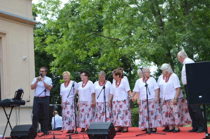 Na scenie stoją kobiety w jednakowych strojach. Po obu stronach kobiet stoją dwaj mężczyźni. Mężczyzna mówi do mikrofonu.