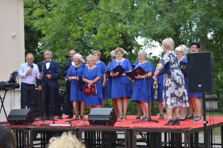 Na scenie stoją mężczyźni i kobiety. Śpiewają. W tle drzewa.