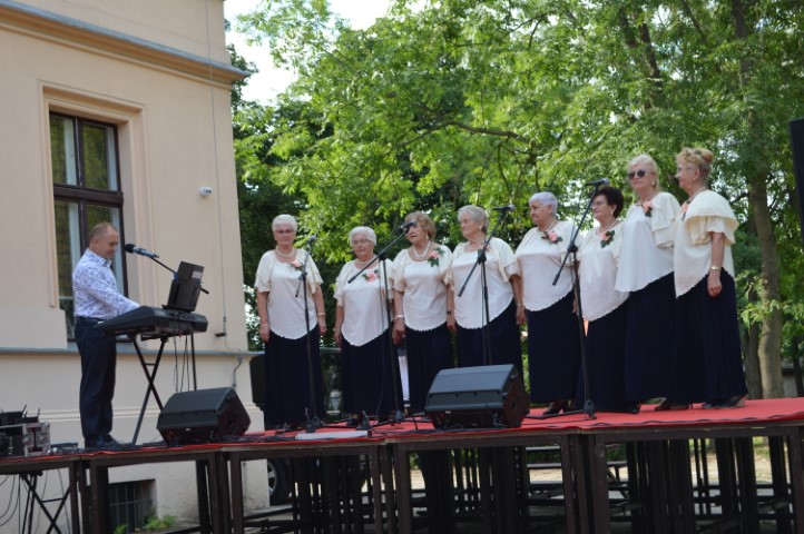 Na scenie w plenerze stoją kobiety ubrane w jednakowe stroje: długie, czarne spódnice, jasne bluzki. Z boku stoi mężczyzna przy instrumencie klawiszowym. W tle drzewa.