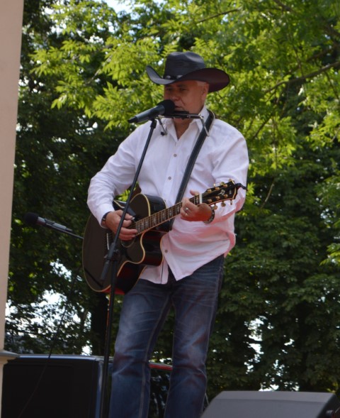 Na scenie w plenerze stoi mężczyzna z gitarą. Na głowie ma kapelusz.
