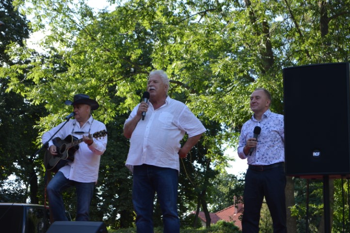 Na scenie w plenerze stoją trzej mężczyźni. Dwaj trzymają w dłoni mikrofony. Jeden gra na gitarze.