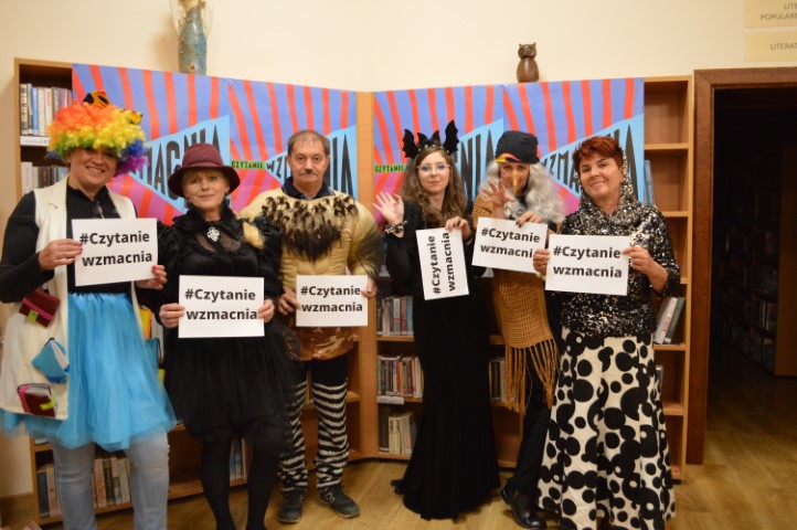 Pracownicy biblioteki w przebraniu pozują na tle ścianki z plakatami z napisem Czytanie Wzmacnia. Trzymają w dłoniach kartki z hasłem Czytanie Wzmacnia