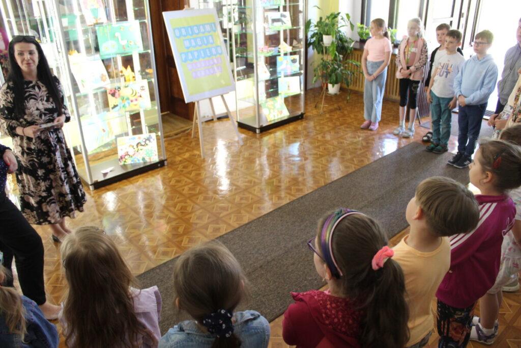 Wystawa prac konkursowych „Origami magia papieru - wiosenne podróże”
zdjęcie grupowe, uczestnicy konkursu 
16 dzieci i 2 dorosłe kobiety, w budynku na drugim planie wystawa prac