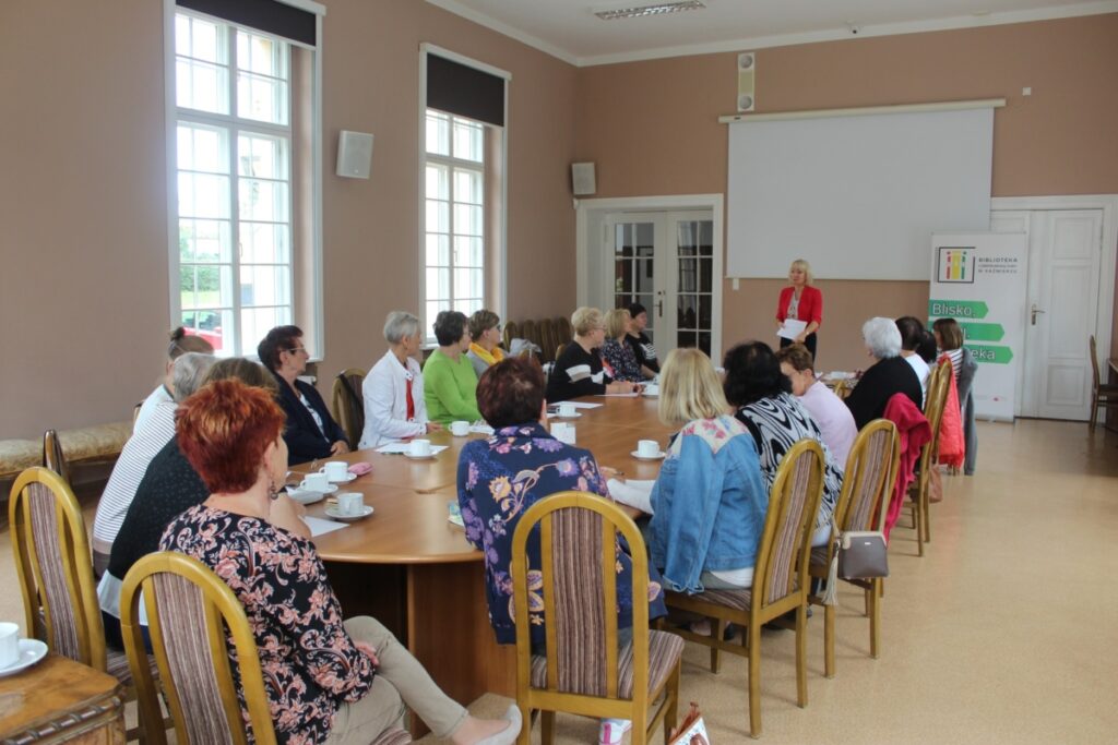 Grupa osób, 20 kobiet siedzi przy stole słucha wykładu jedna prowadząca kobieta