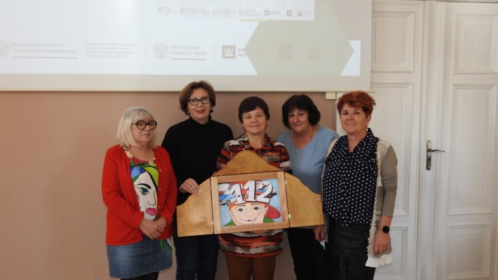 Pięć kobiet, kobieta na środku, trzyma  skrzynkę kamishibai z obrazkiem chłopca i numerem 112
w tle plansza Blisko 