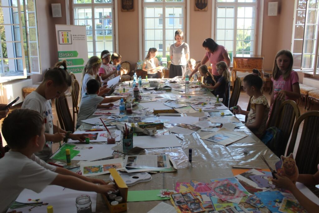 Grupa dzieci siedzi przy stole dorosła kobieta stoi przy stole w tle baner Blisko Bliżej Biblioteka 8.09.2023