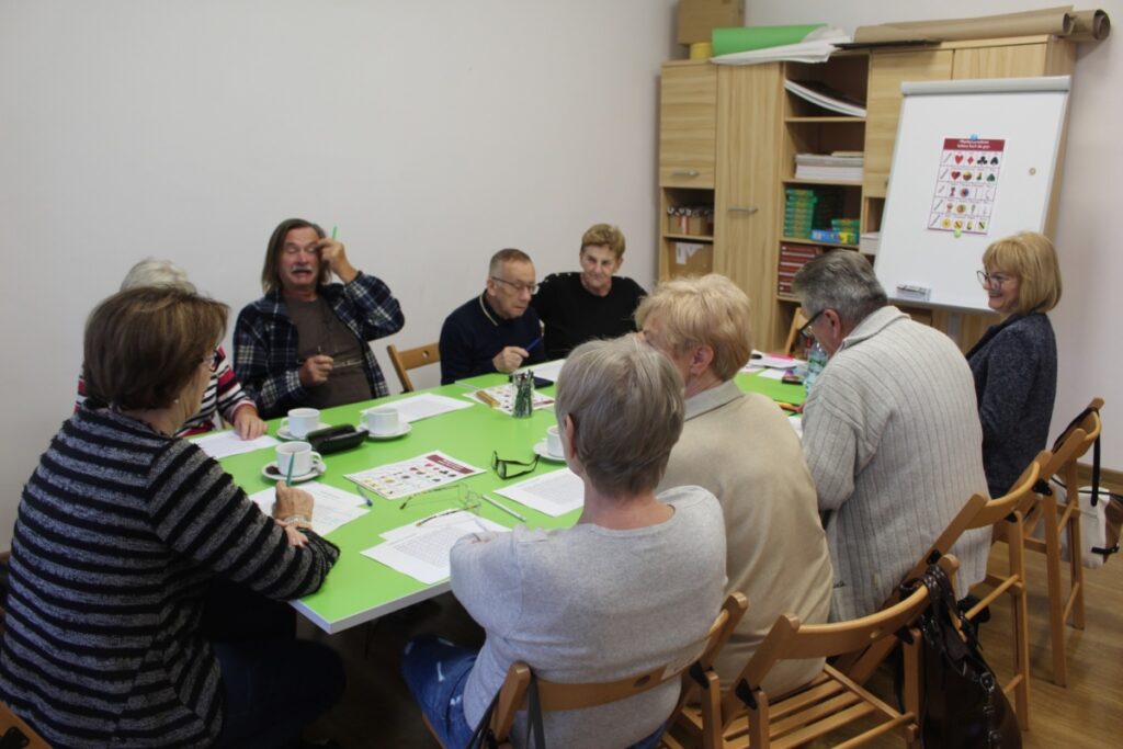 osiem osób w budynku siedzi przy stole Aktywny Senior Zawsze Młody czyli trening umysłu.