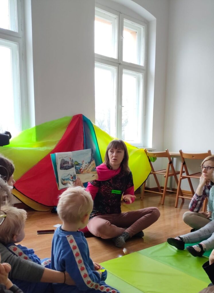 Kobieta siedzi na podłodze pokazuje dzieciom książkę 