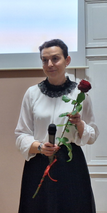 kobieta stoi w budynku, w ręku trzyma mikrofon i różę
