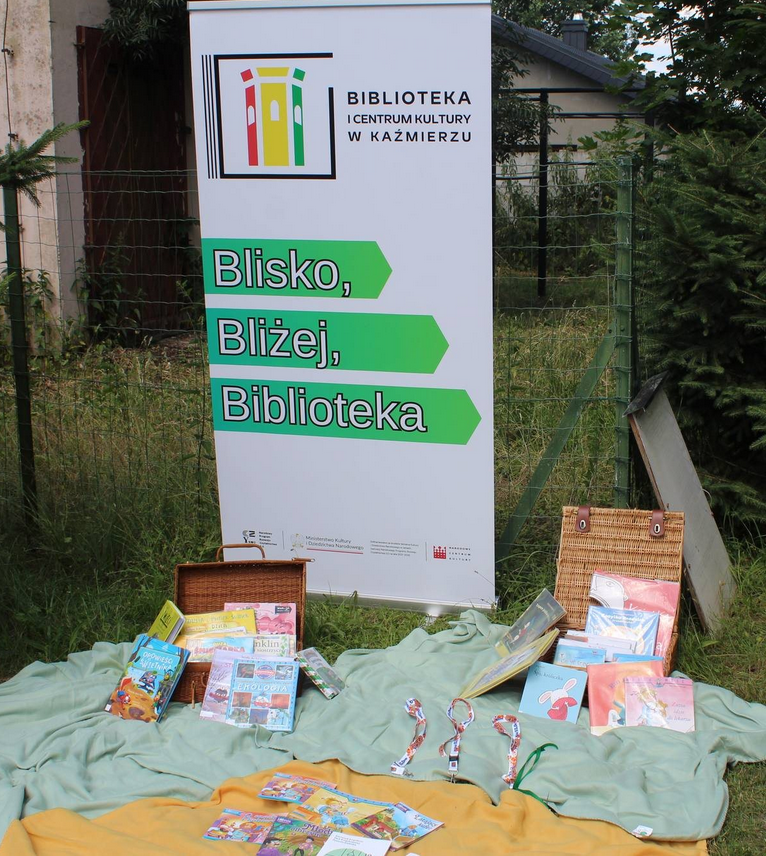 baner blisko bliżej biblioteka nprcz2.0 na powietrzu, koce na trawie na nich książki logo biblioteki w Kaźmierzu