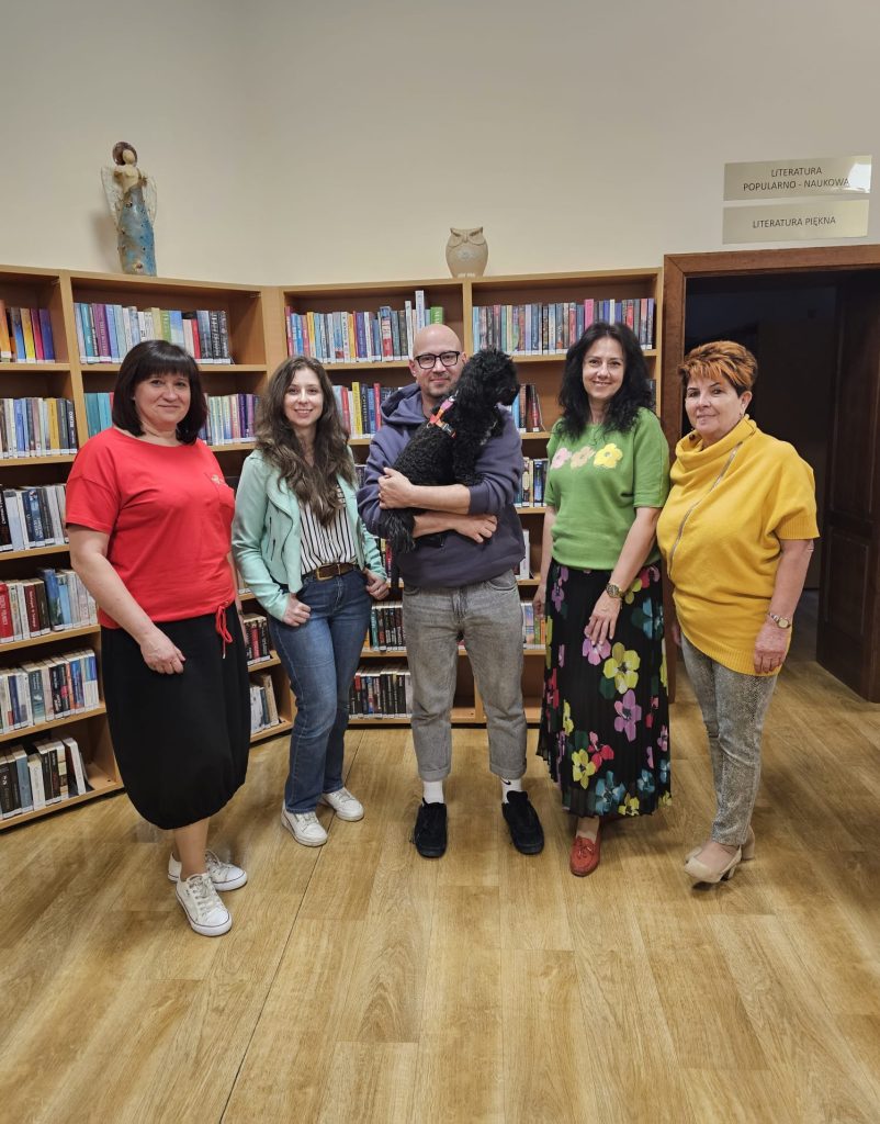 Na tle książek w bibliotece grupa ludzi 1 mężczyzna i 4 kobiety. mężczyzna (stoi na środku) trzymający psa - Marcin Margielewski.