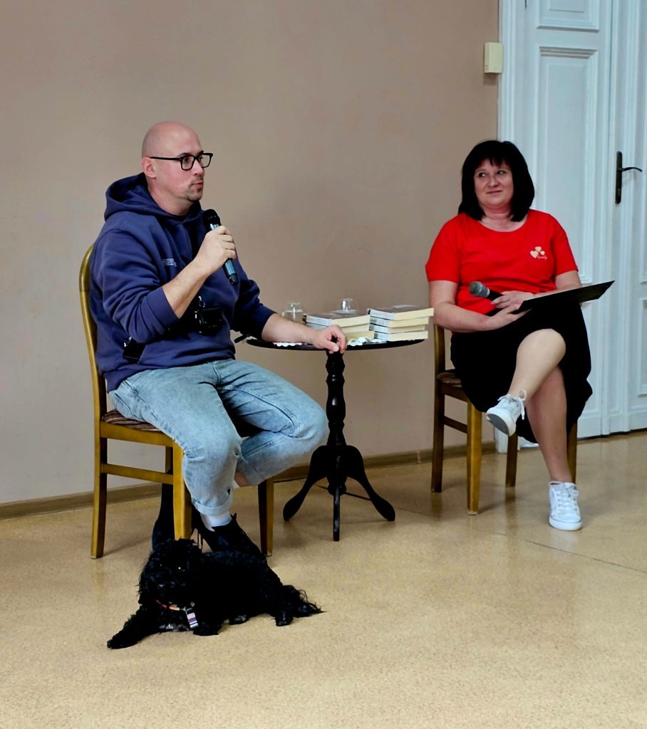 Mężczyzna i kobieta siedzą przy stoliku rozmawiają, trzymają mikrofony , pies leży obok mężczyzny
