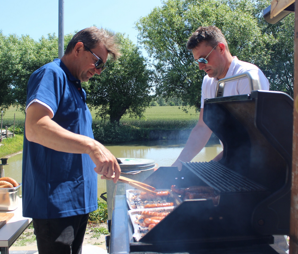 dwóch mężczyzn stoi przy grillu, jeden z nich smaży kiełbasy