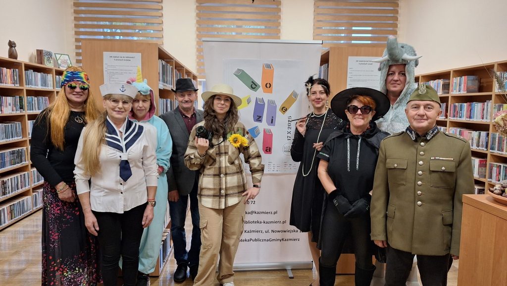 8 osób w pomieszczeniu pozuje do zdjęcia na tle plakatu Noc Bibliotek Do dzieła