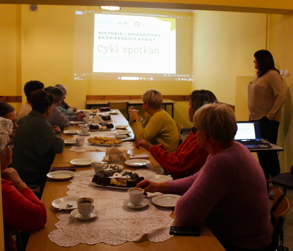spotkanie dwanaście osób przy stole jedna osoba stoi przed prezentacją 
wyświetlana prezentacja z rzutnika 