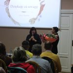 kobieta przemawia do siedzącej grupy ludzi w tle prezentacja „Historia i Dziedzictwo Kaźmierskich Kobiet”