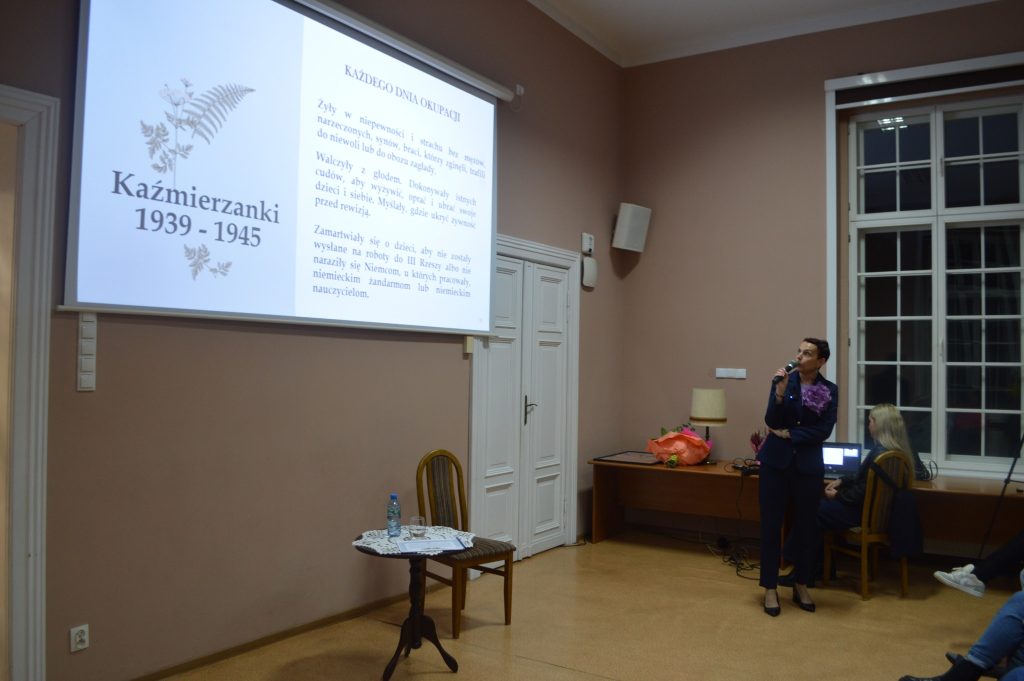 kobieta przemawia do siedzącej grupy ludzi w tle prezentacja „Historia i Dziedzictwo Kaźmierskich Kobiet”