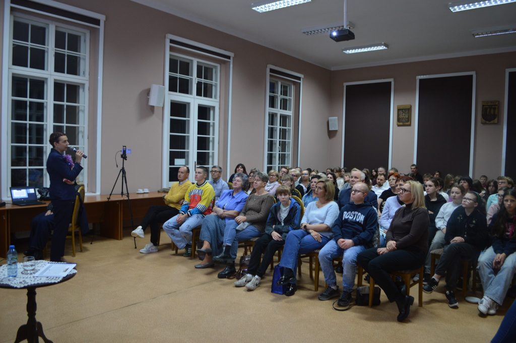 kobieta przemawia do siedzącej grupy ludzi w tle prezentacja „Historia i Dziedzictwo Kaźmierskich Kobiet”