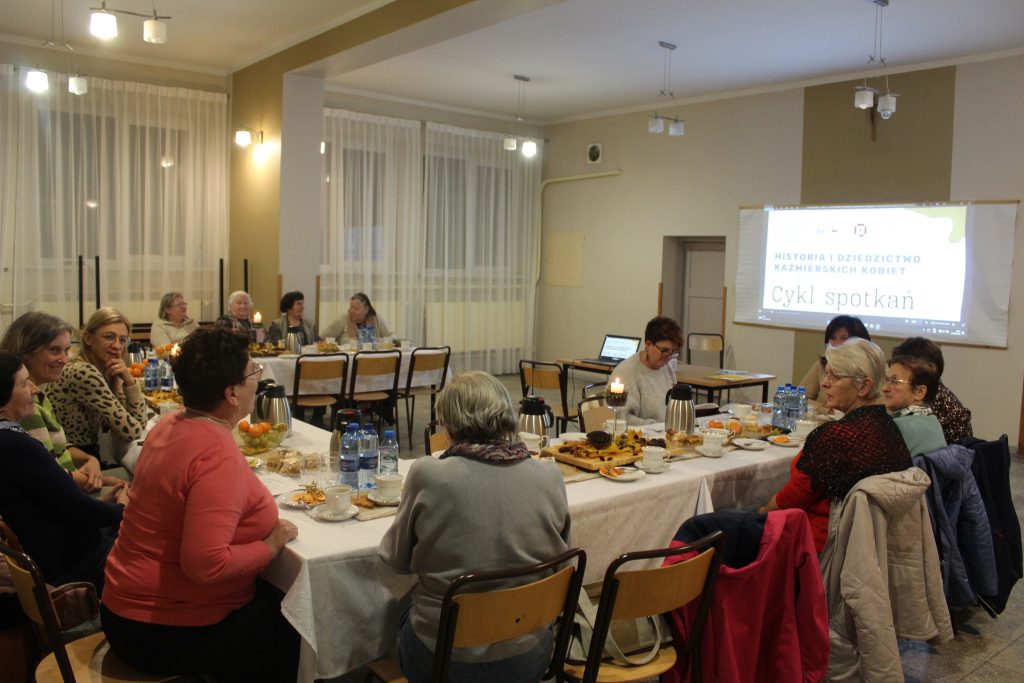 12 osób siedzi przy nakrytym stole ustawionym w podkowę na rzutniku wyświetlana prezentacja