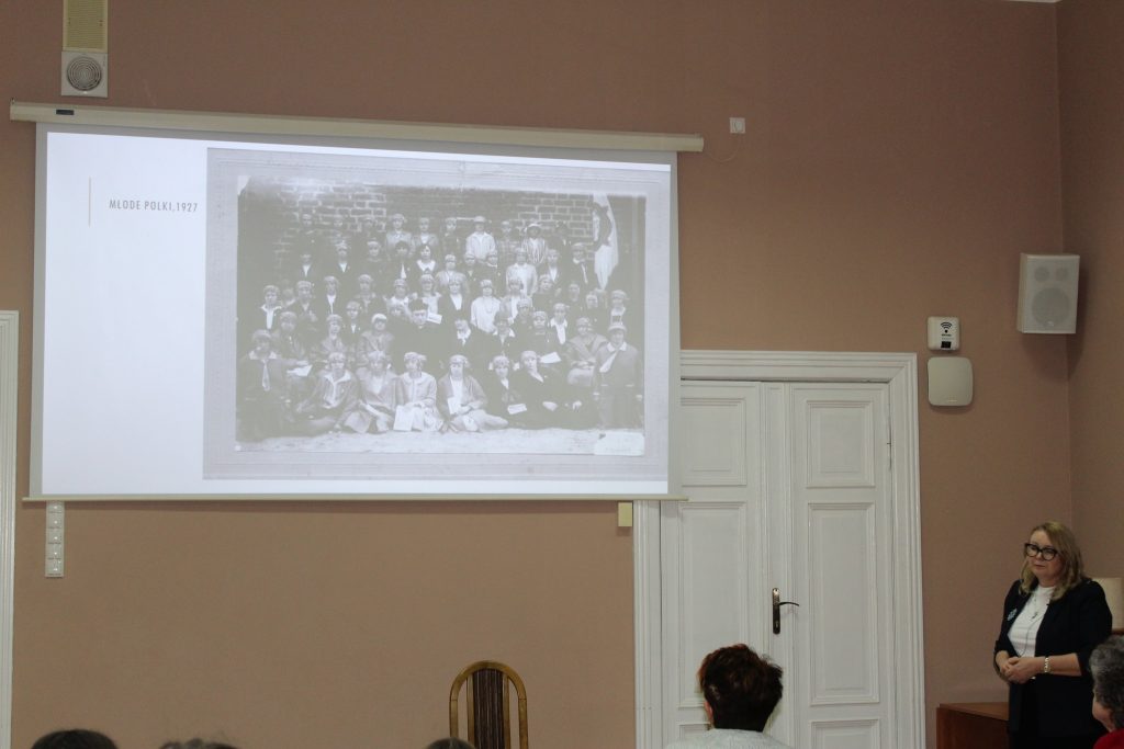 kobieta przemawia do siedzącej grupy ludzi w tle prezentacja „Historia i Dziedzictwo Kaźmierskich Kobiet”