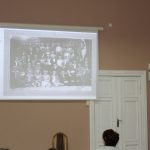 kobieta przemawia do siedzącej grupy ludzi w tle prezentacja „Historia i Dziedzictwo Kaźmierskich Kobiet”