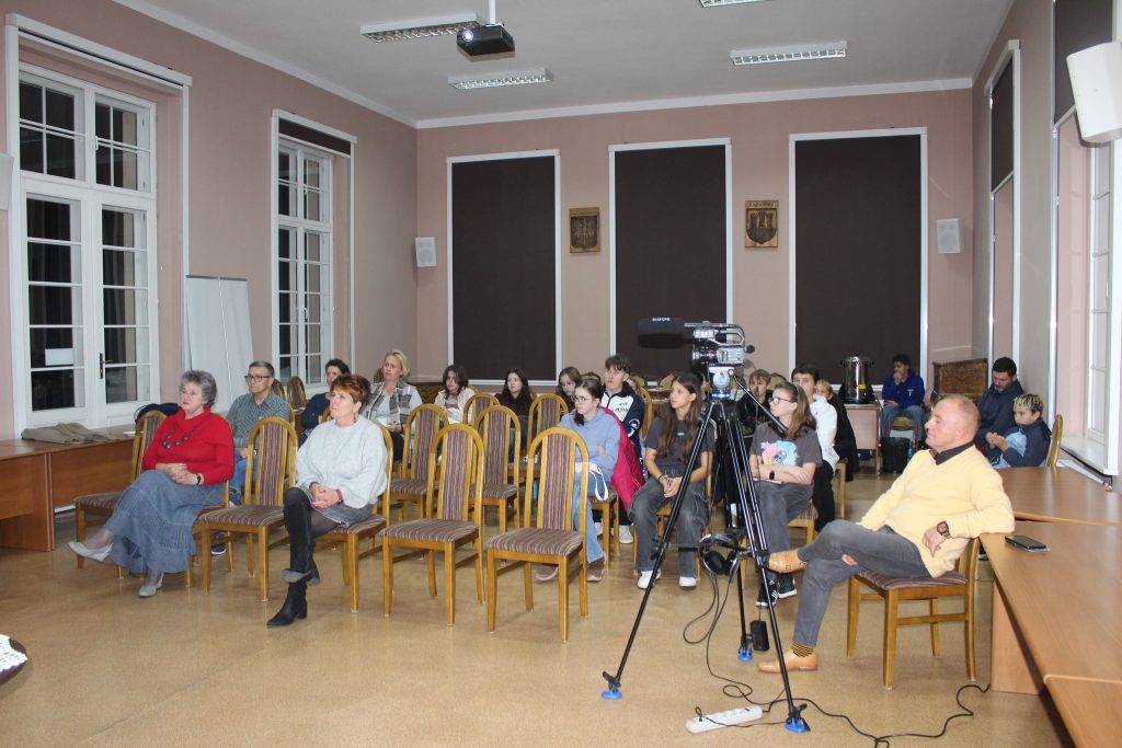 kobieta przemawia do siedzącej grupy ludzi w tle prezentacja „Historia i Dziedzictwo Kaźmierskich Kobiet”