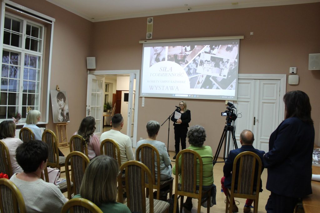 kobieta przemawia do siedzącej grupy ludzi w tle prezentacja „Historia i Dziedzictwo Kaźmierskich Kobiet” „Siła i codzienność. Kobiety Gminy Kaźmierz”