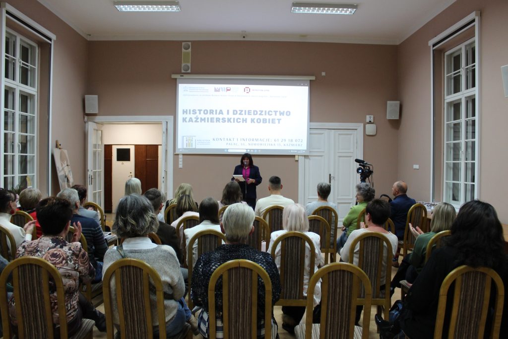 kobieta przemawia do siedzącej grupy ludzi w tle prezentacja „Historia i Dziedzictwo Kaźmierskich Kobiet”