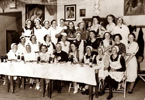 Kurs dorabiania wina 1938
grupa kobiet i ksiądz
część kobiet siedzi za stołami długi stół przykryty obrusem na nim wina
część kobiet stoi na drugim planie