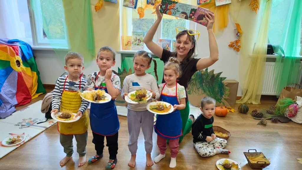 Kolorowy pokuj kobieta na 2 planie pokazuje książkę uśmiecha się 5 dzieci pokazuje swoje prace z warsztatów sensorycznych