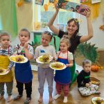 Kolorowy pokuj kobieta na 2 planie pokazuje książkę uśmiecha się 5 dzieci pokazuje swoje prace z warsztatów sensorycznych