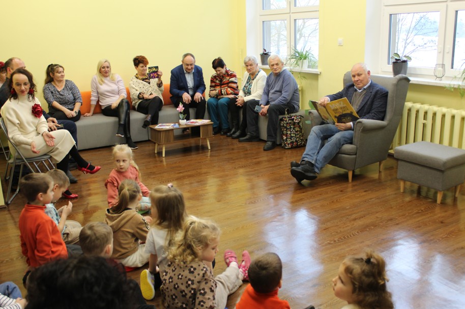 grupa ludzi w pomieszczeniu dzieci i dorośli mężczyzna czyta dzieciom książkę 