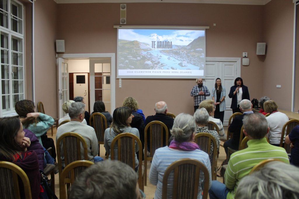 W budynku prezentacja ludzie siedzą 3 osoby stoją jedna osoba kobieta przemawia przez mikrofon