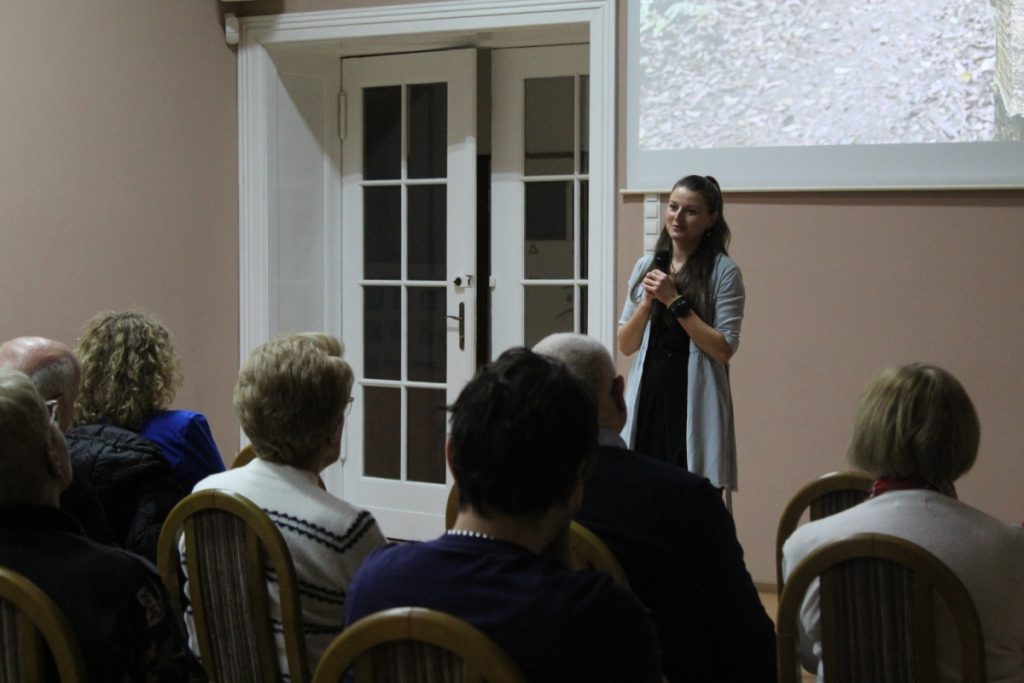 W budynku prezentacja ludzie siedzą jedna osoba kobieta przemawia przez mikrofon