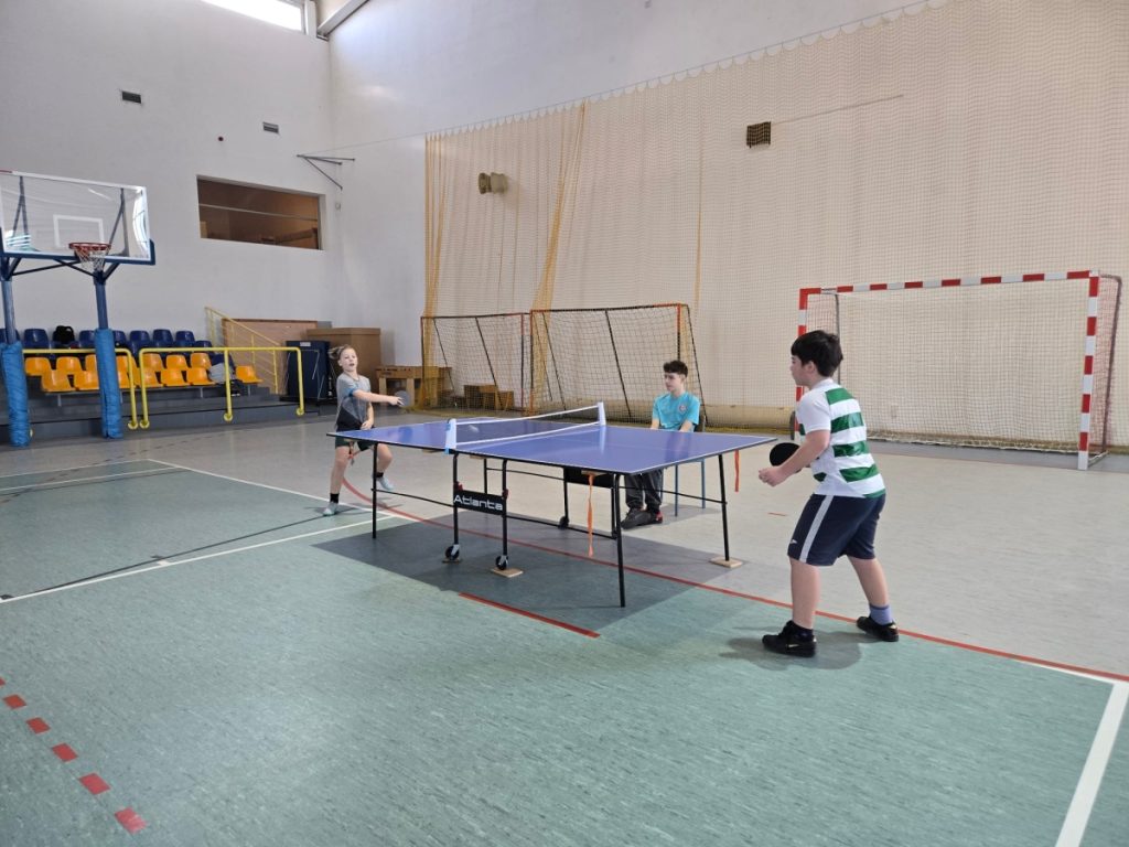 dwóch chłopców gra w tenisa stołowego na hali sportowej jeden chłopiec siedzi przy stole i sędziuje 