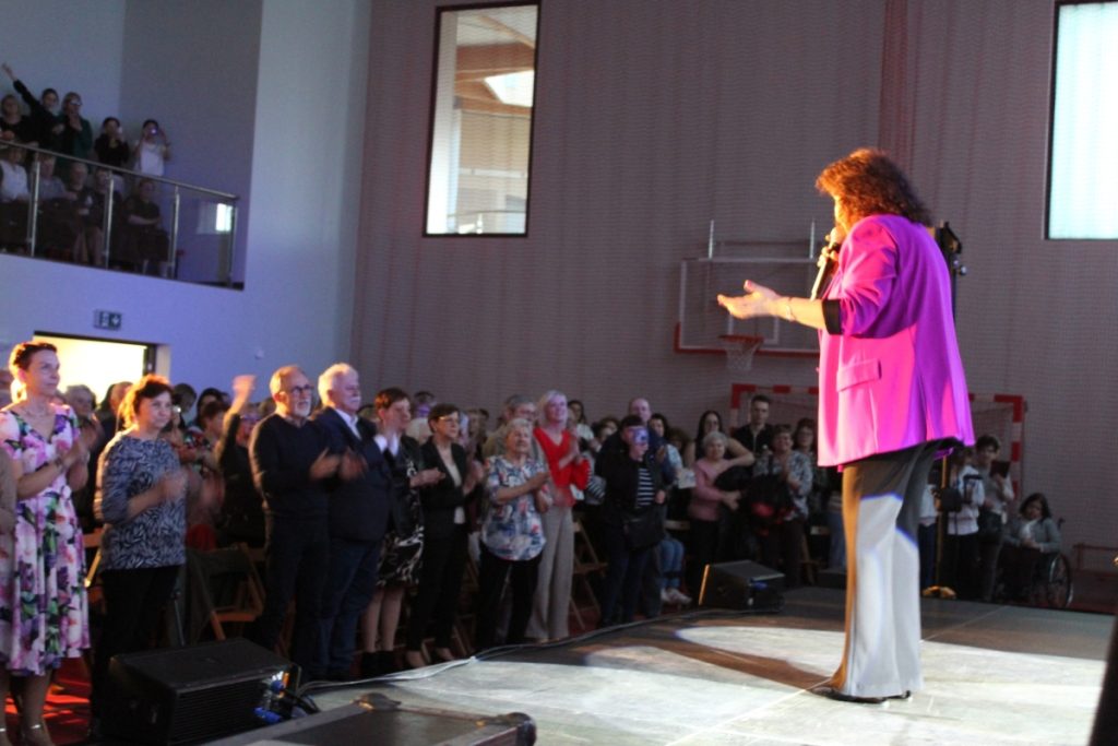 w pomieszczeniu, na scenie śpiewa piosenkarka, publiczność stoi i klaszcze 