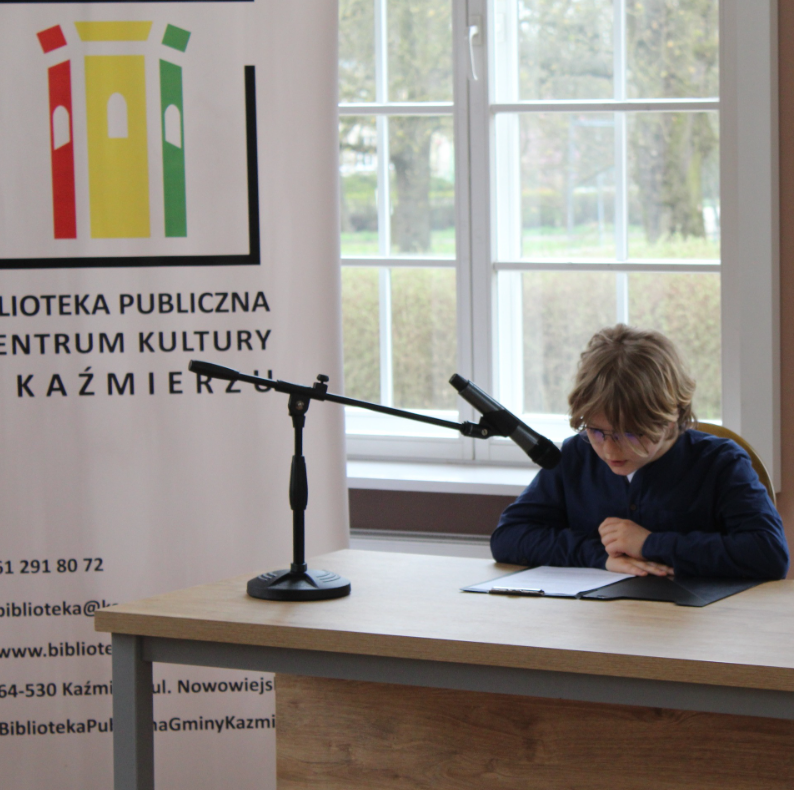 chłopiec czyta do mikrofonu, siedzi przy stole, czyta, za nim logo biblioteki