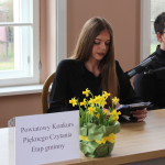 dziewczyna siedzi przy stole, kwiaty, napis: Powiatowy konkurs Pięknego czytania etap gminny