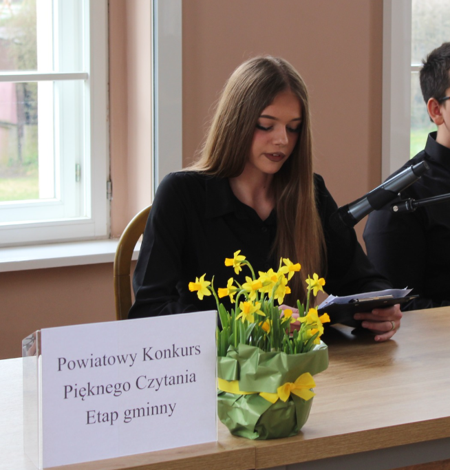 dziewczyna siedzi przy stole, kwiaty, napis: Powiatowy konkurs Pięknego czytania etap gminny 