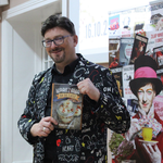 autor książek Alek Rogoziński, pozuje z książką, w budynku, w tle baner